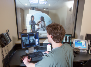 Interior of a room containing MRI machine. One healthcare staff reviews scans, another helps a patient on the MRI table.