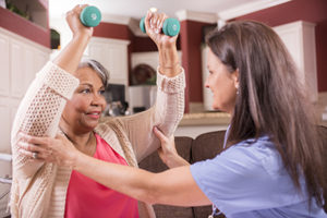 Older woman holds small weights above her head with help from healthcare staff.
