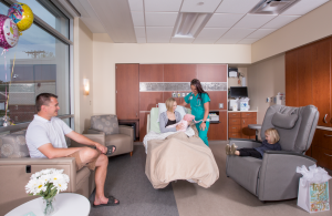 Hospital room. Mother sitting in a bed holding a new born baby next to a nurse. Adult male and toddler sit on the furniture.