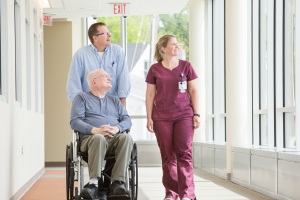 An older man in a wheelchair is being pushed by an adult male, while female healthcare worker stands beside them.