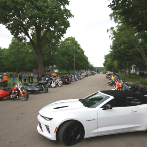 Motorcycles line both sides of a street along with a white sports car.