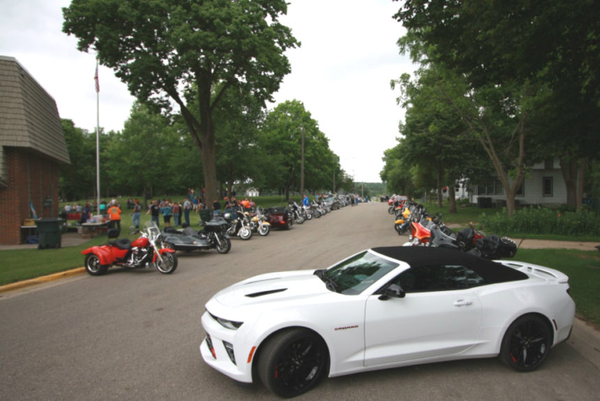 Motorcycles line both sides of a street along with a white sports car.