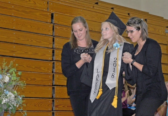 Young woman in cap and gown, wearing a sash that reads “Lakers 2018” walks to graduate from high school, with help