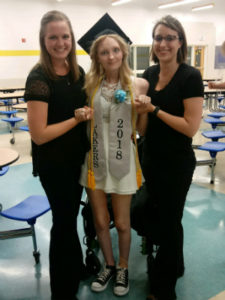 Young woman in cap and gown with sash that reads “Lakers 2018” stands next to two female healthcare staff.