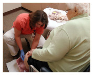 Female healthcare staff member kneels to wash feet of older woman.
