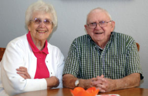 married white hair couple with glasses, sitting at kitchen table, white wall, smiling