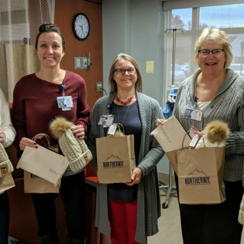 Five women in hospital room holding brown paper gift bags and cream colored winter hats.