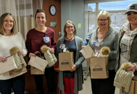 Five women in hospital room holding brown paper gift bags and cream colored winter hats.