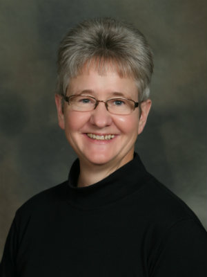 LaNita Mortenson, PT, Physical Therapist, studio picture of older caucasian woman smiling, short grey hair and glasses, black shirt